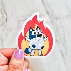 May include: A die-cut vinyl sticker featuring a cartoon blue heeler dog with dark blue patches and a white muzzle, standing in front of a stylized red and yellow flame graphic. The dog has a determined expression with raised fists.