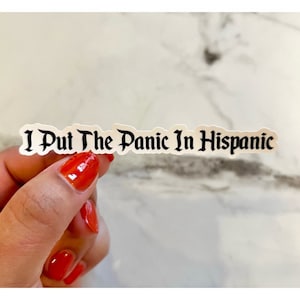 May include: A white sticker with the phrase "I Put The Panic In Hispanic" in black gothic font. The sticker is held by a hand with red nail polish.