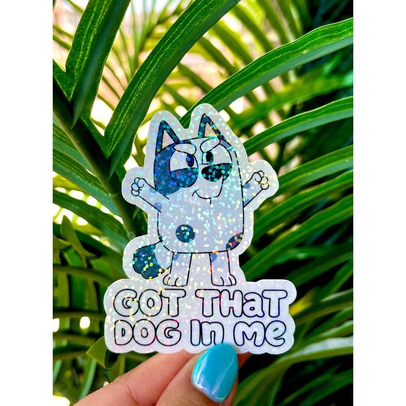 May include: A holographic sticker featuring a cartoon blue heeler dog with a determined expression, standing with arms raised. The sticker includes the text "GOT THAT DOG IN ME" in bold, rounded black lettering against a shimmering background.