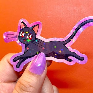 Könnte beinhalten: Ein gestanzter Aufkleber einer schwarzen Katze mit großen roten Augen und einem gelben Halbmond auf der Stirn. Die Katze ist in einer Sprungpose dargestellt und hat ein holografisches Finish mit verstreuten bunten Sternen und einem rosa Rand.