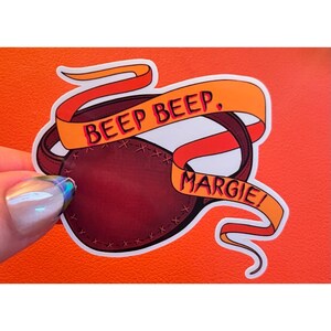 K&ouml;nnte beinhalten: Ein gestanzter Aufkleber mit einer dunkelbraunen Augenklappe in Lederoptik und sichtbaren N&auml;hten an den R&auml;ndern. Ein orangefarbenes Band windet sich um die Augenklappe mit dem Text BEEP BEEP, MARGIE! in fetten roten Gro&szlig;buchstaben.