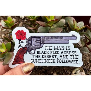 Könnte beinhalten: Ein gestanzter Aufkleber mit einer detaillierten Illustration eines Revolvers mit einem verzierten roten Griff und einer roten Rose, die sich um den Griff windet. Der Text lautet, The man in black fled across the desert, and the gunslinger followed.