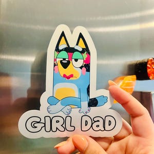 May include: A die-cut vinyl sticker featuring a cartoon blue dog character with a tired expression, wearing a pink headband and sitting cross-legged. Below the character, the text GIRL DAD is printed in bold, black, rounded capital letters.