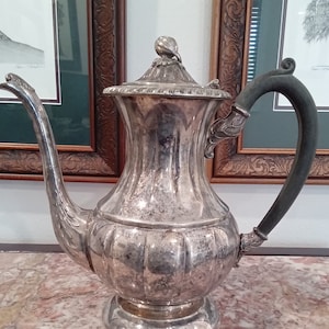 May include: A silver teapot with a black handle and a decorative lid. The teapot is sitting on a marble surface.