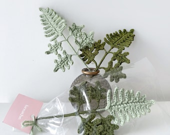 May include: A bouquet of crocheted green fern leaves in a clear glass vase with a brown textured surface. The vase is sitting on a white surface. The bouquet is packaged in a clear plastic bag with a pink tag that says "breathe."