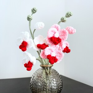 May include: A bouquet of crocheted pink and white orchids with red centres in a textured grey glass vase.