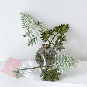 May include: A bouquet of crocheted green fern leaves in a clear glass vase with a brown textured surface. The vase is sitting on a white surface. The bouquet is packaged in a clear plastic bag with a pink tag that says "breathe."