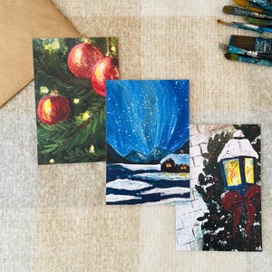 Juego de 3 impresiones artísticas de postales navideñas de invierno pintadas a mano: acogedora cabaña navideña, paisaje nevado, linterna navideña y adornos brillantes.