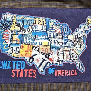 May include: A navy blue patch featuring a map of the United States, composed of license plates. The patch is sewn onto a plaid shirt. The text "UNITED STATES OF AMERICA" is printed below the map in blue and red.