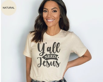 May include: A woman wearing a cream colored t-shirt with the text "Y'all Need Jesus" printed in black. The shirt has a short sleeve and a crew neck.