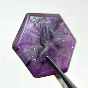 Puede incluir: Una piedra preciosa púrpura con inclusiones negras, cortada en forma hexagonal. La piedra preciosa está sujeta por unas pinzas.