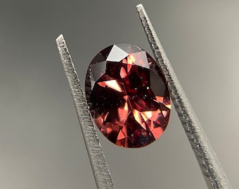 May include: A red-brown oval gemstone with a faceted cut, held by tweezers against a gray background.