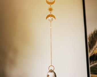 May include: A gold chain with three crescent moon charms and a sun charm hangs from a crystal ball. The chain is suspended from a hook.