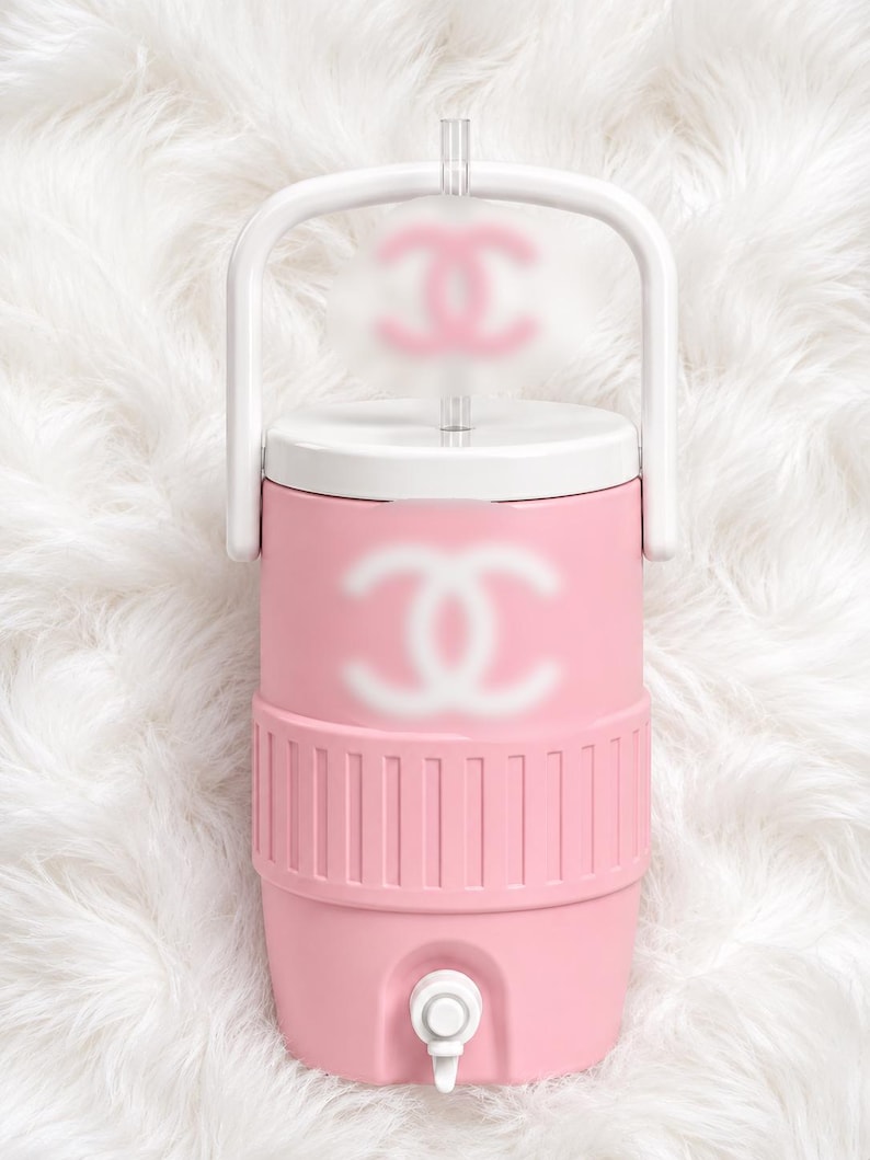 May include: A pink water cooler with a white handle and lid. The cooler features a white logo on the front and top. It has a spigot at the bottom and a clear straw. The cooler is sitting on a white, fluffy surface.