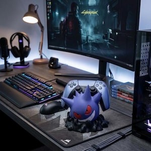 May include: A gaming setup featuring a curved monitor displaying a video game scene. A purple Gengar figure sits on a mousepad, with a controller behind it. A keyboard, mouse, headphones, and a desk lamp complete the setup.