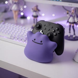 May include: A black Nintendo Switch controller rests on a purple Ditto-shaped stand. The stand has a smiling face. The background includes a computer monitor, keyboard, and figurines, all in a purple and white color scheme.