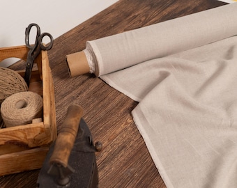 May include: A still-life image featuring craft supplies on a wooden surface. A roll of light beige fabric sits next to a wooden crate filled with twine and scissors. An antique iron is also present.