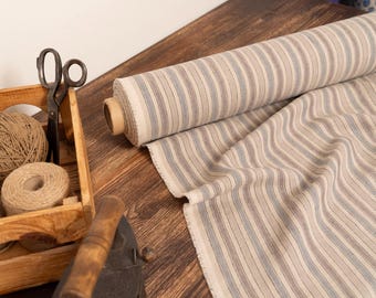 Può includere: Un rotolo di tessuto a righe nei toni beige, grigio e blu, accanto a una cassa di legno piena di spago e rocchetti, e un paio di forbici vintage. Un ferro da stiro antico poggia su una superficie di legno.