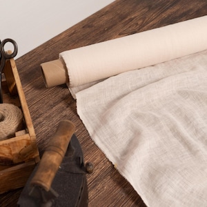 May include: A wooden crate contains spools of twine and vintage scissors. A roll of off-white fabric sits next to a piece of unrolled fabric. An antique iron is in the foreground.