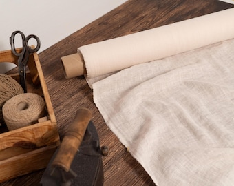 Puede incluir: Una caja de madera contiene carretes de hilo y unas tijeras antiguas. Un rollo de tela blanquecina se encuentra junto a un trozo de tela desenrollada. Una plancha antigua está en primer plano.
