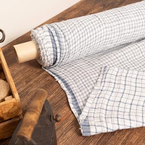 Könnte beinhalten: Eine Rolle blau-weiß karierter Stoff liegt auf einer Holzoberfläche. Eine Holzkiste enthält Schnur und eine Schere. Ein antikes Bügeleisen ist ebenfalls vorhanden, was auf ein Näh- oder Bastelthema hindeutet.