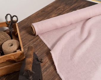 May include: A roll of light pink fabric sits on a wooden surface next to a wooden box filled with twine spools and scissors. An antique iron is in the foreground. The scene suggests a craft or sewing project.