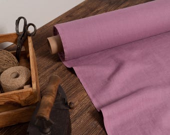 May include: A roll of dusty rose fabric sits on a wooden surface, with a wooden crate containing twine and scissors. An antique iron rests nearby. The scene evokes a vintage craft or sewing theme.