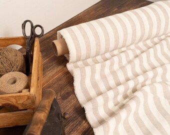 May include: A roll of beige and white striped fabric sits on a wooden surface, alongside a wooden crate filled with twine and scissors. The fabric is partially unrolled, revealing its pattern.
