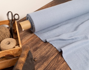 May include: A roll of light blue fabric sits on a wooden surface, with a wooden crate containing twine and scissors. The fabric is unrolled, showing its texture. An antique iron is also visible.