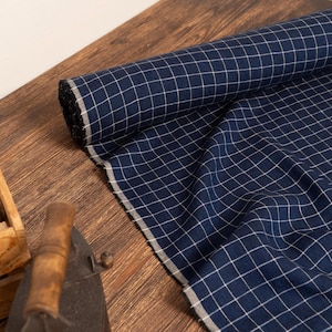 May include: A roll of navy blue fabric with a white grid pattern is displayed on a wooden surface. A wooden box with scissors, twine, and an iron are also visible. The fabric is partially unrolled, showing its texture.