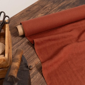 Puede incluir: Un rollo de tela color óxido se despliega sobre una superficie de madera. Una caja de madera contiene hilo y carretes, con tijeras vintage. Una plancha antigua descansa cerca, sugiriendo un tema de artesanía o costura.