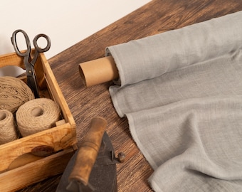 May include: A wooden crate holds twine balls and scissors, next to a roll of gray fabric. An antique iron rests nearby. The scene suggests a craft or sewing project, with natural materials and tools.