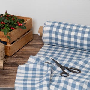 May include: A roll of blue and white checkered fabric sits next to a pair of black scissors. A wooden crate holds a plant with green leaves and red berries. A spool of twine is also present.