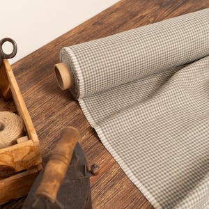 May include: A roll of gray and white checkered fabric sits on a wooden surface, with a portion unrolled to the side. A wooden crate holds twine and scissors, and an antique iron rests nearby. The scene suggests a craft or sewing theme.