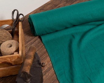 Op de afbeelding: Een rol teal stof met een houten kist met touw en een schaar. Een antieke strijkijzer staat in de buurt. De stof is uitgespreid op een houten oppervlak, wat wijst op een knutsel- of naaiproject.