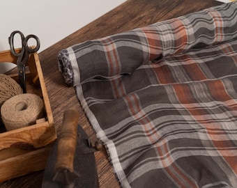 May include: A roll of gray, orange, and white plaid fabric lies on a wooden surface. A wooden box holds twine, scissors, and a metal tool. The fabric is partially unrolled, revealing the pattern.