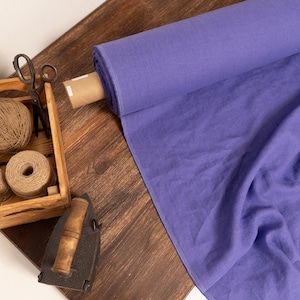 Può includere: Un rotolo di tessuto viola con una cassa di legno contenente spago e forbici. Un ferro da stiro antico è vicino. Il tessuto è di una vibrante tonalità viola, e la scena è su una superficie di legno.