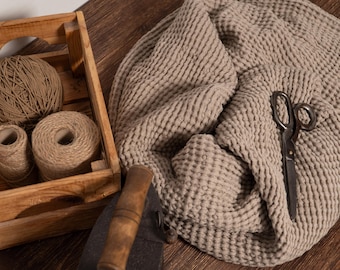Pode incluir: Uma caixa de madeira contém barbante e bobinas de fio de cor natural. Um tecido de waffle bege grande e texturizado é drapeado sobre uma superfície de madeira, com um par de tesouras vintage em cima. Uma alça de madeira é visível.