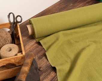 May include: A wooden crate holds twine, thread spools, and vintage scissors. A roll of olive green fabric sits next to the crate, with some fabric unrolled. The items are on a wooden surface, suggesting a craft or sewing theme.