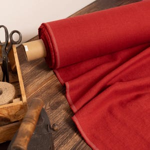 Könnte beinhalten: Eine Rolle leuchtend roter Stoff liegt auf einer Holzoberfläche, ein Teil ist ausgerollt, um seine Textur zu zeigen. Eine Holzkiste enthält Schnur und eine Schere, und ein antikes Bügeleisen liegt daneben, was auf ein Näh- oder Bastelthema hindeutet.