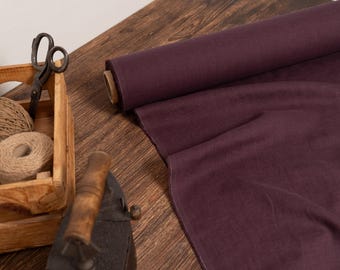 May include: A roll of deep purple fabric sits on a wooden surface, with a portion unrolled. A wooden crate holds twine and scissors. An antique iron is in the foreground. The scene suggests a craft or sewing theme.