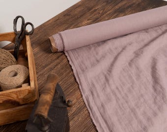 Puede incluir: Un rollo de tela rosa polvoriento con un borde deshilachado, junto a una caja de madera llena de hilo y tijeras. Una plancha antigua descansa cerca sobre una superficie de madera, lo que sugiere un tema de artesanía o costura.