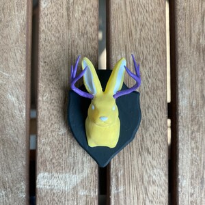 May include: A yellow rabbit with purple antlers mounted on a black plaque. The rabbit has white ears and a white nose.