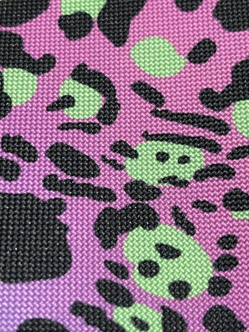 May include: Purple fabric with a black and green abstract pattern. The pattern resembles a series of overlapping circles and swirls.