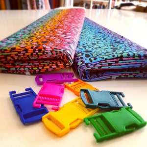 May include: A close-up of colorful plastic clasps or buckles, some are blue, green, yellow, pink, and purple. The clasps are on a white surface next to two folded pieces of fabric with a leopard print pattern. One fabric is a rainbow gradient and the other is a blue gradient.
