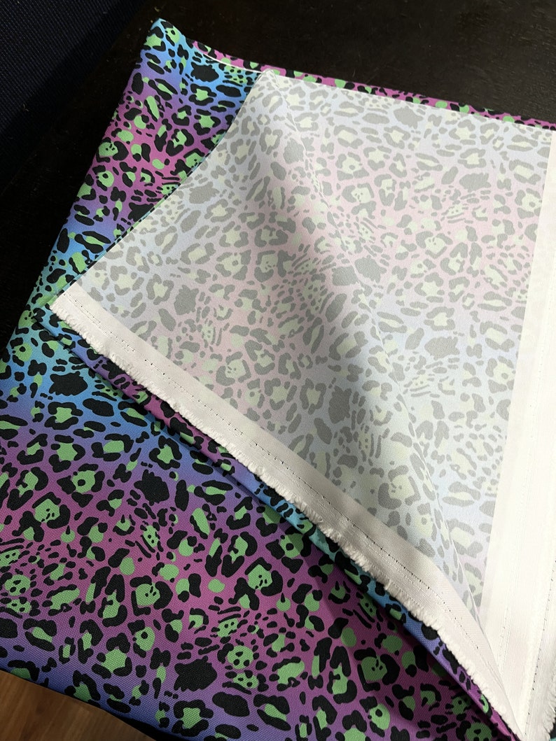 May include: A close-up of a fabric with a leopard print pattern in shades of blue, purple, green, and black. The fabric is folded over, revealing a white lining.