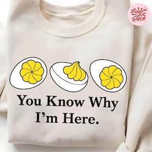 Può includere: Felpa color crema con grafica di tre uova ripiene. Le uova hanno ripieno giallo e sono delineate in nero. Il testo "You Know Why I'm Here" è stampato in nero sotto. Il logo Popcorn Art Gallery è in alto a destra.