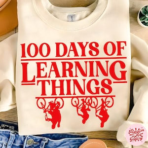May include: Cream-colored sweatshirt with the text "100 DAYS OF LEARNING THINGS" in red. Below the text are three red silhouettes of children riding bicycles. The sweatshirt is folded on a wooden surface.