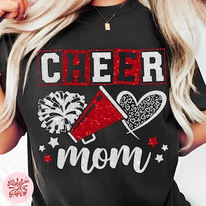 Op de afbeelding: Zwart t-shirt met een rood en wit ontwerp. Het ontwerp bevat het woord "CHEER" in een rood omlijnde doos, een megafoon, een pompon, een hart en het woord "mom". Witte sterren en rode accenten maken het ontwerp compleet.