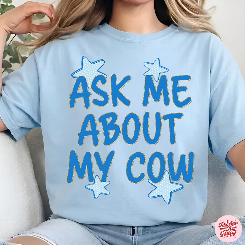 May include: Light blue t-shirt with the text "ASK ME ABOUT MY COW" in blue, outlined in gold. Four blue stars with white polka dots are around the text. The shirt is a casual, everyday wear item.